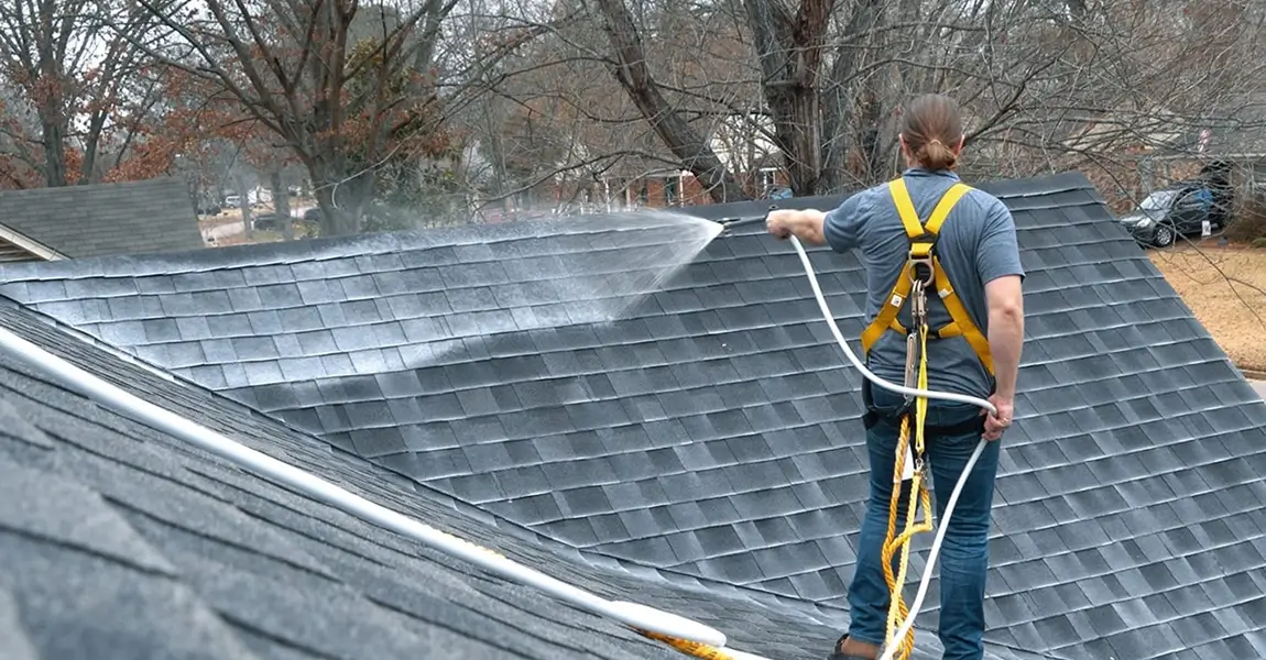 Harnessed worker using a spray wand across an asphalt-shingle roof