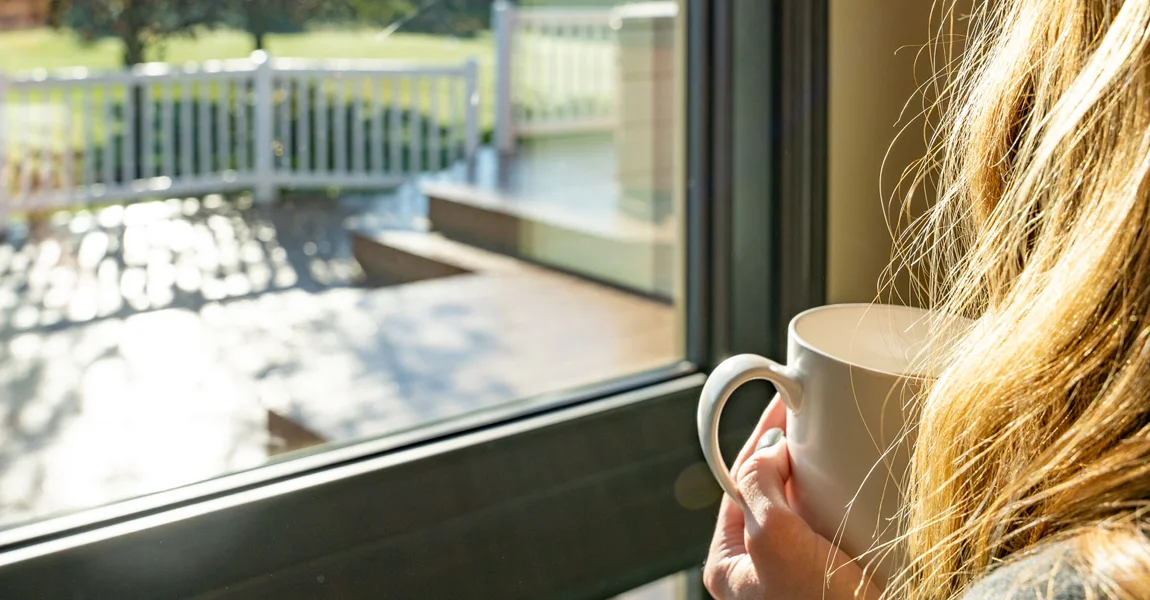 A person holding a cup beside a sunlit window overlooking a yard